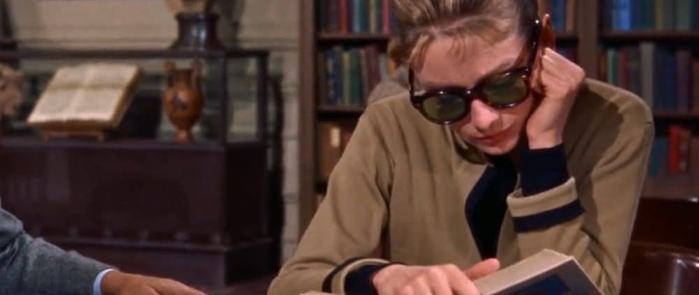 Image of Audrey Hepburn in the library reading a book and wearing sunglasses.