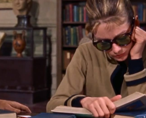 Image of Audrey Hepburn in the library reading a book and wearing sunglasses.