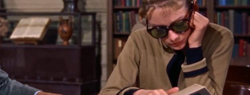 Image of Audrey Hepburn in the library reading a book and wearing sunglasses.