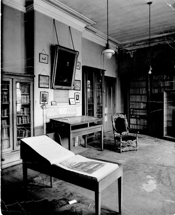 A historical black-and-white photograph of an antique library study room featuring slanted book lecterns, glass cases, tall bookcases, and a large portrait hanging above a fireplace.