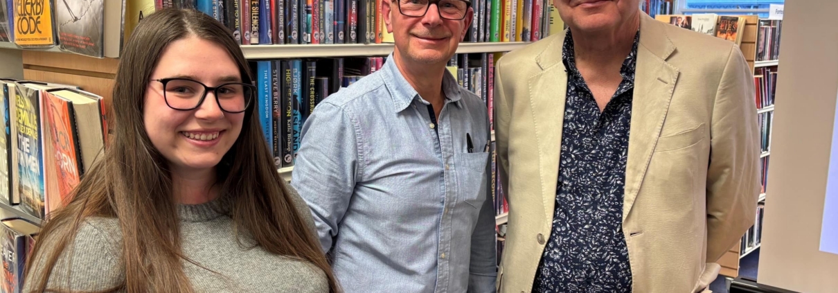 Gibson Library Society Secretary, Chairman and Treasurer standing together in front of bookshelves filled with fiction novels.