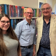 Gibson Library Society Secretary, Chairman and Treasurer standing together in front of bookshelves filled with fiction novels.