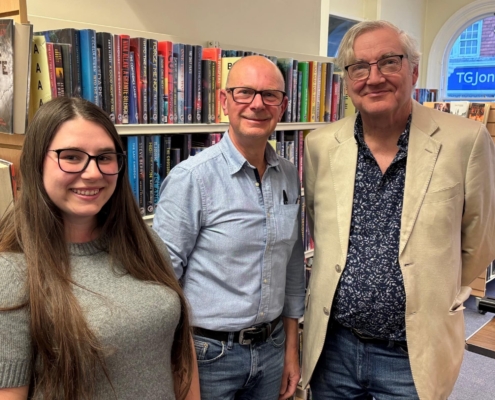 Gibson Library Society Secretary, Chairman and Treasurer standing together in front of bookshelves filled with fiction novels.