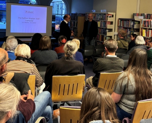 An audience seated in a library watching a presentation by sculptor Ian Wolter titled "The Saffron Walden 1647 memorial."