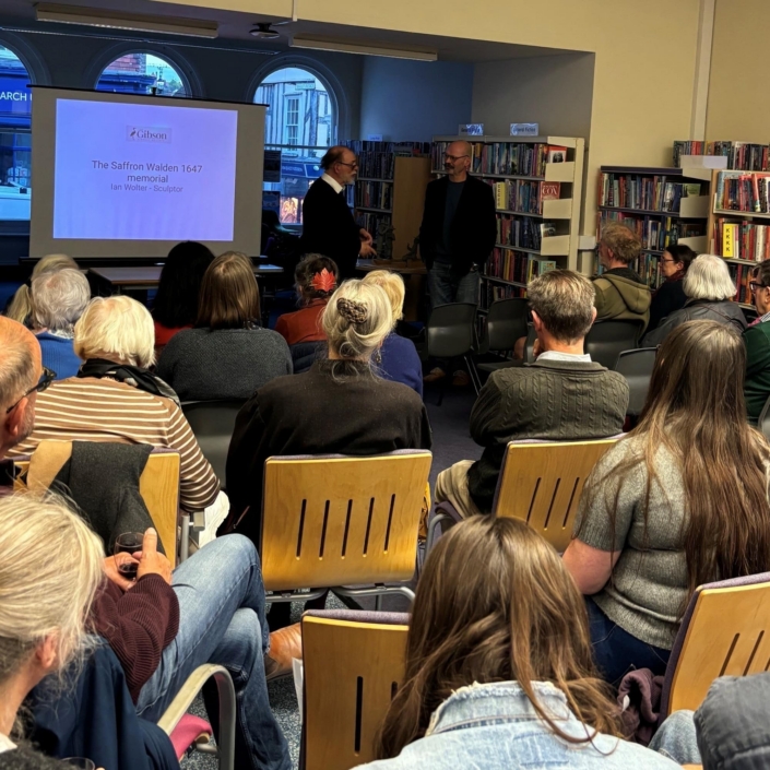An audience seated in a library watching a presentation by sculptor Ian Wolter titled "The Saffron Walden 1647 memorial."