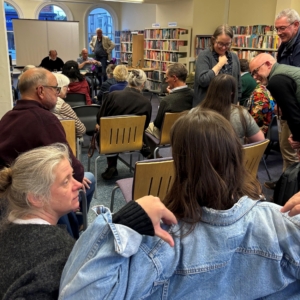 An AGM gathering inside a library, showing rows of seated audience members and groups socialising between bookshelves.