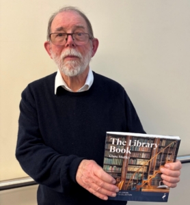 A photograph of a man (Martyn Everett) with a white beard and glasses holding a copy of "The Library Book" by Clare Mulley against a plain wall.