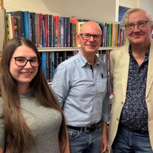 Gibson Library Society Secretary, Chairman and Treasurer standing together in front of bookshelves filled with fiction novels.
