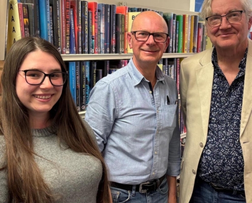 Gibson Library Society Secretary, Chairman and Treasurer standing together in front of bookshelves filled with fiction novels.