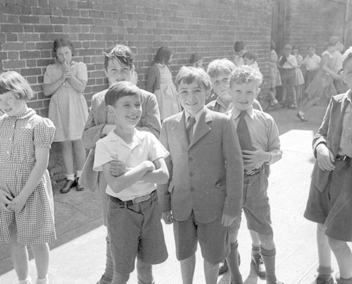 A mid-20th-century black-and-white photograph of school children in traditional dresses and short-trouser suits posing in a brick courtyard.