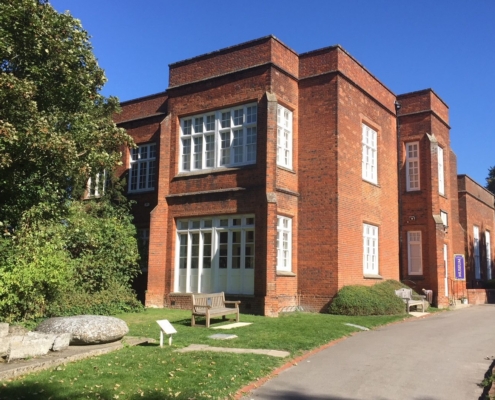 The red-brick exterior of the Saffron Walden Museum with white multi-paned windows, a paved pathway, and a wooden bench on the lawn.