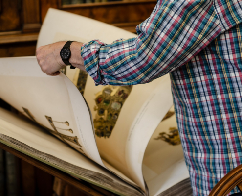 A close-up of a person turning the large, illustrated pages of an antique volume resting on a wooden reading desk.