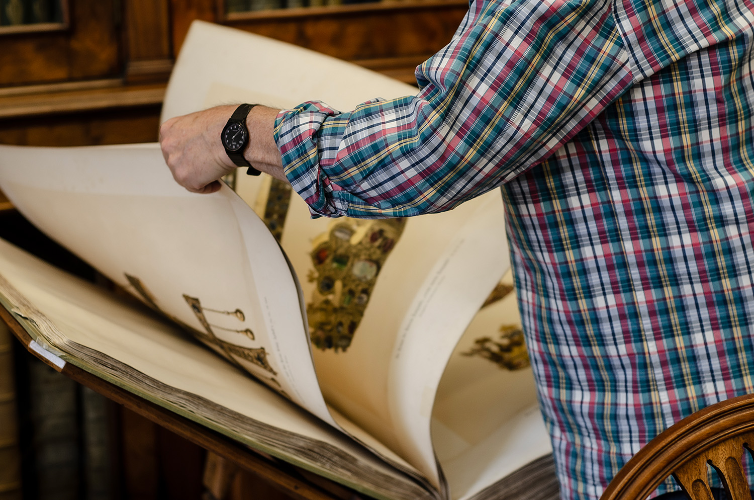 A close-up of a person turning the large, illustrated pages of an antique volume resting on a wooden reading desk.